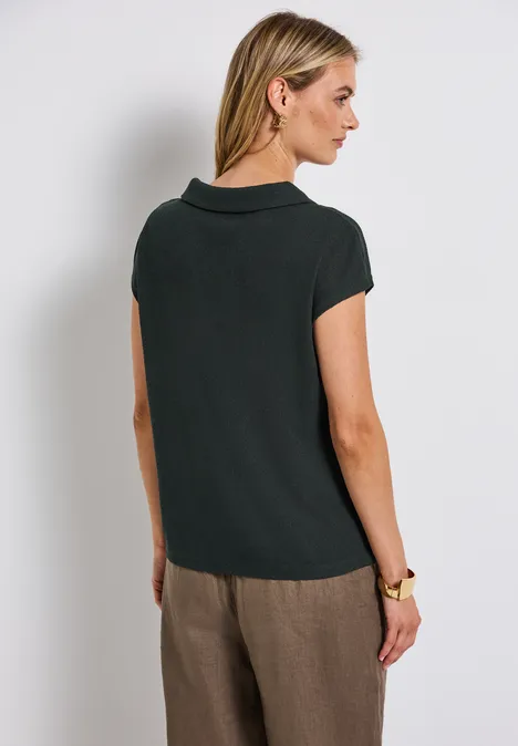 Frau mit dunkelgrünem Shirt und brauner Hose steht seitlich, Blick nach rechts.